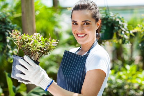 A young woman with dark hair tied back, wearing a white t-shirt, a blue striped apron, and gardening gloves, is smiling while holding a potted shrub with small green leaves and pinkish edges in an outdoor garden setting. The background features lush green foliage, wooden garden structures, and vibrant plants, indicating a well-maintained landscaped garden with a variety of plants, including a hedge and flower beds, under bright natural light on a clear day. The scene suggests outdoor gardening activities such as planting or pruning, typical of professional gardening services in the Seven Kings area, part of London Borough of Redbridge, with the focus on garden maintenance and planting.
